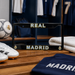 A black license plate frame with 'REAL MADRID' in gold and white lettering, displayed on a wooden bench in a Real Madrid locker room setting with soccer shoes, a soccer ball, and a navy jersey nearby.