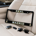 A black license plate frame with 'REAL MADRID' in white and yellow lettering, displayed on a beige leather car seat next to car keys and sunglasses.