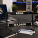A black license plate frame with 'REAL MADRID' in white and gold lettering, displayed on a dark wooden desk with various Real Madrid themed items in the background.