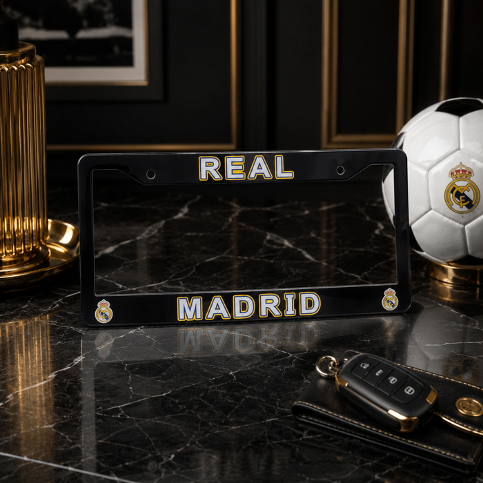 A black license plate frame featuring 'REAL MADRID' in gold and white lettering with the club's crest, displayed on a dark marble surface next to a soccer ball and car key fob.