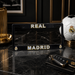 A black license plate frame featuring 'REAL MADRID' in gold and white lettering with the club's crest, displayed on a dark marble surface next to a soccer ball and car key fob.