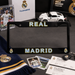 A black Real Madrid license plate frame displayed on a dark tabletop with team merchandise around it, including a scarf, cards, tickets, a model car, and branded boxes.