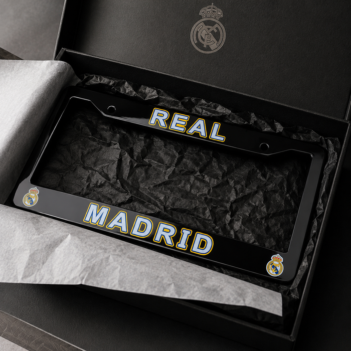 A black Real Madrid license plate frame with 'REAL' on the top bar and 'MADRID' on the bottom bar, featuring the club's crest on each corner. The frame is presented inside an open black gift box lined with tissue paper.
