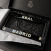 A black Real Madrid license plate frame with 'REAL' on the top bar and 'MADRID' on the bottom bar, featuring the club's crest on each corner. The frame is presented inside an open black gift box lined with tissue paper.