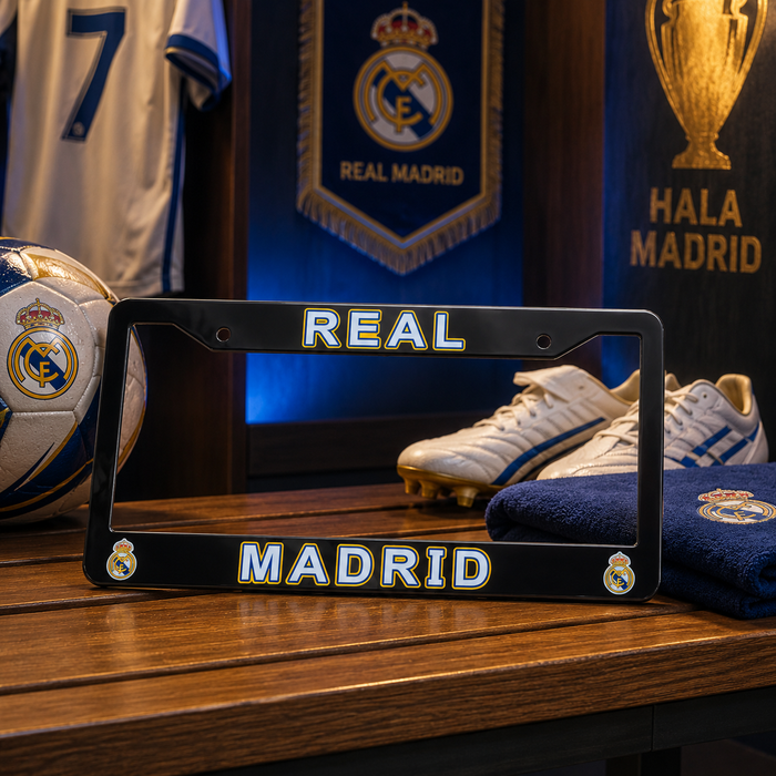 A black Real Madrid license plate frame with gold and white lettering sits on a wooden table, surrounded by soccer-themed items including a Real Madrid soccer ball, white cleats, a blue towel, and jerseys in the background.