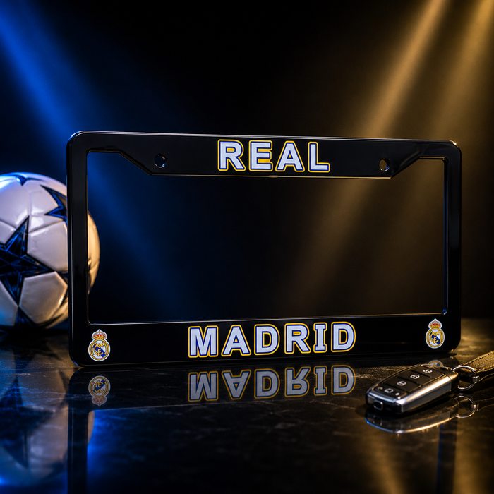 A black license plate frame with 'REAL MADRID' in white letters outlined in gold, featuring the Real Madrid crest in the corners, displayed on a reflective dark surface with a soccer ball and car key in the background.