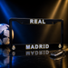 A black license plate frame with 'REAL MADRID' in white letters outlined in gold, featuring the Real Madrid crest in the corners, displayed on a reflective dark surface with a soccer ball and car key in the background.