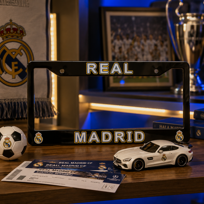 A black Real Madrid license plate frame with gold and white lettering, displayed on a wooden table alongside a miniature white Mercedes-AMG sports car, a soccer ball, a ticket, and a toy car, with a blurred Real Madrid interior background.