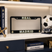 A black license plate frame with 'REAL MADRID' in white and gold lettering, displayed on a white shelf next to a soccer ball, books, and a model car.