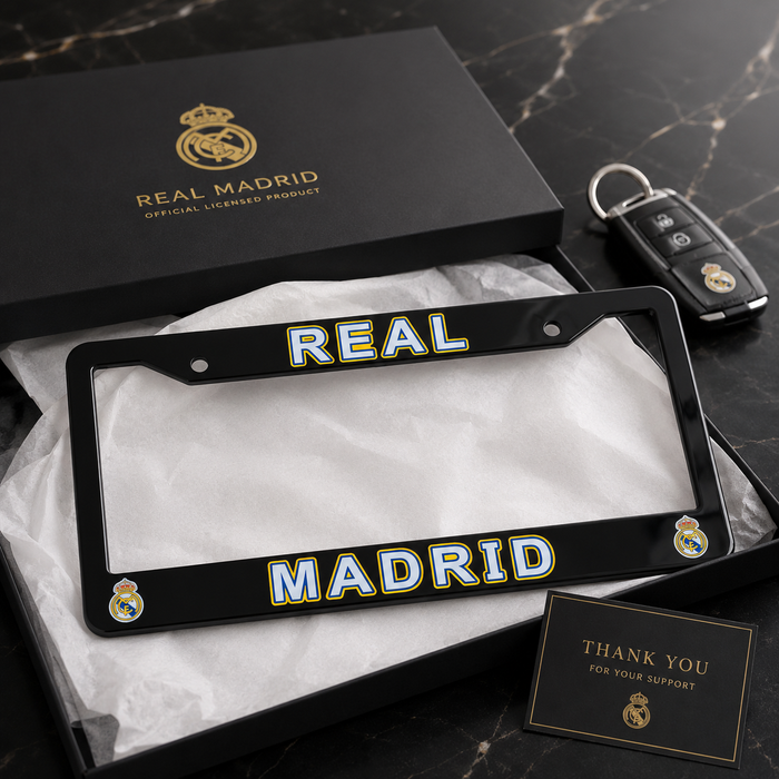 A black Real Madrid license plate frame with 'REAL MADRID' in white and gold lettering, displayed in an open black presentation box with tissue paper. The Real Madrid branded box lid and a car key are visible behind it, with a small thank-you card in front, all on a dark marble surface.