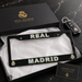 A black Real Madrid license plate frame with 'REAL MADRID' in white and gold lettering, displayed in an open black presentation box with tissue paper. The Real Madrid branded box lid and a car key are visible behind it, with a small thank-you card in front, all on a dark marble surface.