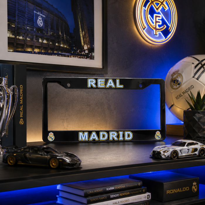 A black license plate frame with 'REAL MADRID' in blue LED-style lettering, displayed on a shelf with Real Madrid-themed decor including a soccer ball, framed stadium photo, and model cars.