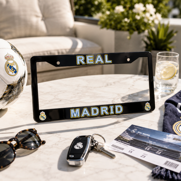 A black license plate frame with 'REAL MADRID' in blue and gold lettering, styled on a marble table with a Real Madrid soccer ball, sunglasses, keys, a drink, and a ticket.