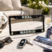 A black license plate frame with 'REAL MADRID' in blue and gold lettering, styled on a marble table with a Real Madrid soccer ball, sunglasses, keys, a drink, and a ticket.