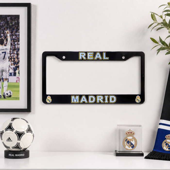 A black license plate frame with 'REAL MADRID' in gold and blue lettering, displayed on a white wall above a shelf with soccer-themed decor, including a framed photo, a soccer ball, a crest display, and a scarf.