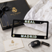 A black Real Madrid license plate frame with 'REAL MADRID' in white and gold lettering, displayed on a marble surface with a branded black gift box, tissue paper, thank you card, and car key.