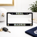 A black license plate frame with 'REAL MADRID' in gold and blue lettering, displayed on a white desk with a Real Madrid soccer ball, keys, a plant, and a navy blue scarf in the background.