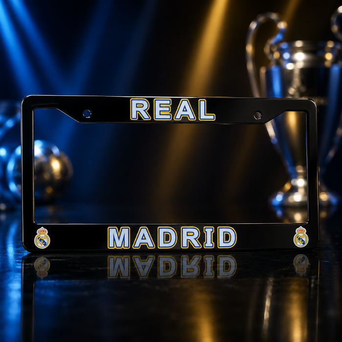 A black license plate frame featuring 'REAL MADRID' in white and gold lettering, with the Real Madrid crest in the bottom corners, displayed on a reflective dark surface with blurred trophies in the background.