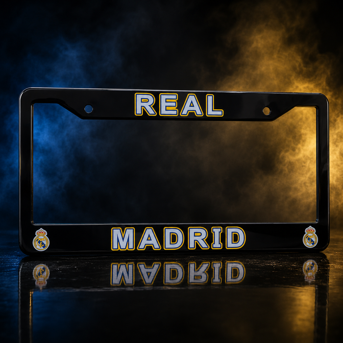 A black Real Madrid license plate frame with 'REAL' at the top and 'MADRID' at the bottom, featuring the club's crest on each corner and a reflective finish, set against a dramatic smoky background with blue and gold lighting.