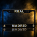 A black Real Madrid license plate frame with 'REAL' at the top and 'MADRID' at the bottom, featuring the club's crest on each corner and a reflective finish, set against a dramatic smoky background with blue and gold lighting.