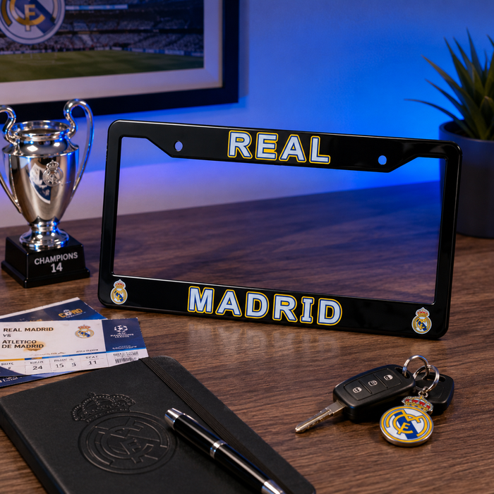 A black Real Madrid license plate frame displayed on a wooden desk, featuring 'REAL MADRID' in white with gold outlines and club crests. The frame is styled with a notebook, pen, keys, ticket, and a silver trophy in the background under blue ambient lighting.