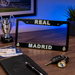 A black Real Madrid license plate frame displayed on a wooden desk, featuring 'REAL MADRID' in white with gold outlines and club crests. The frame is styled with a notebook, pen, keys, ticket, and a silver trophy in the background under blue ambient lighting.