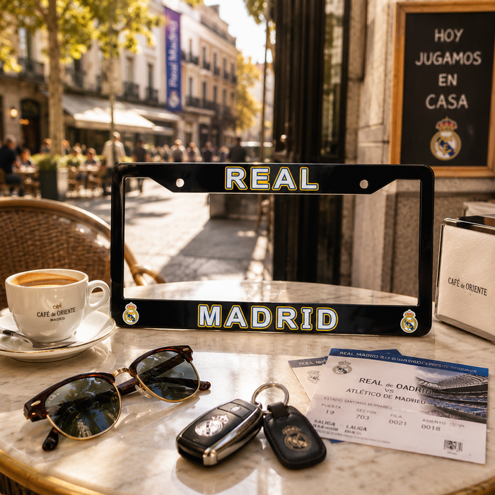 A black license plate frame with 'REAL MADRID' in gold and white lettering, displayed on a cafe table alongside a coffee cup, sunglasses, keys, and tickets, with a blurred street scene in the background.