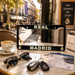 A black license plate frame with 'REAL MADRID' in gold and white lettering, displayed on a cafe table alongside a coffee cup, sunglasses, keys, and tickets, with a blurred street scene in the background.