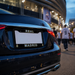 Close-up rear view of a glossy black car with a decorative license plate frame reading "REAL" on top and "MADRID" on the bottom, set in a real outdoor street environment at dusk with blurred people and stadium lights in the background.
