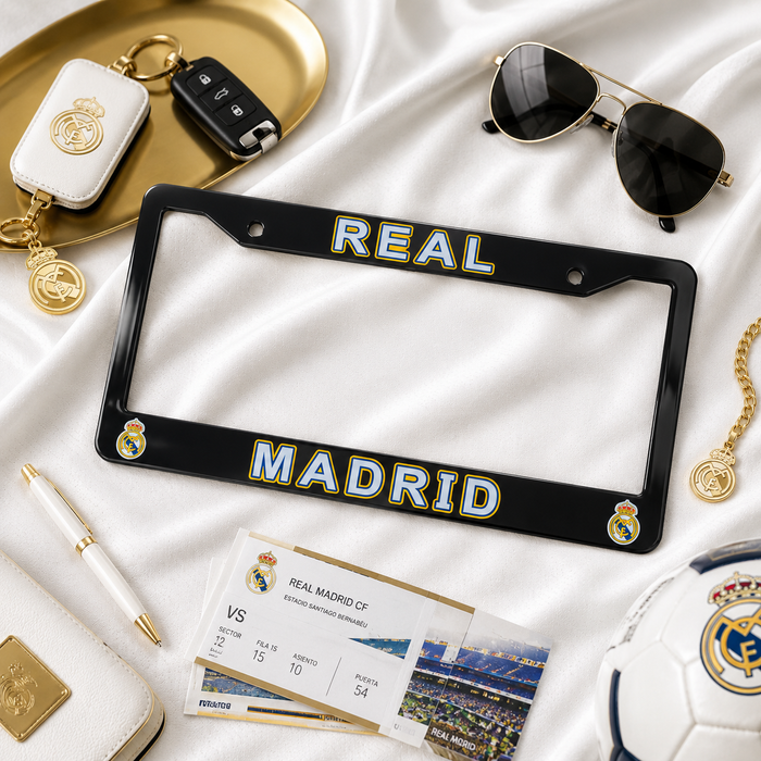 A black license plate frame with 'REAL MADRID' in blue and gold lettering, styled in a flat lay on white satin with Real Madrid-themed accessories including a keychain, sunglasses, tickets, a pen, a wallet, and a soccer ball.