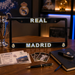 A black Real Madrid license plate frame displayed on a wooden desk, surrounded by football-themed items including a silver trophy, tickets, a notebook, a pen, and a car key fob. The frame features 'REAL MADRID' in white letters with gold and blue outlines, and the club's crest is visible at the bottom corners.