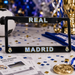 A black Real Madrid license plate frame with 'REAL' at the top and 'MADRID' at the bottom, decorated with gold and blue lettering and small club crests. The frame is displayed on a festive table with confetti, a ticket, a soccer ball, and trophies in the background.