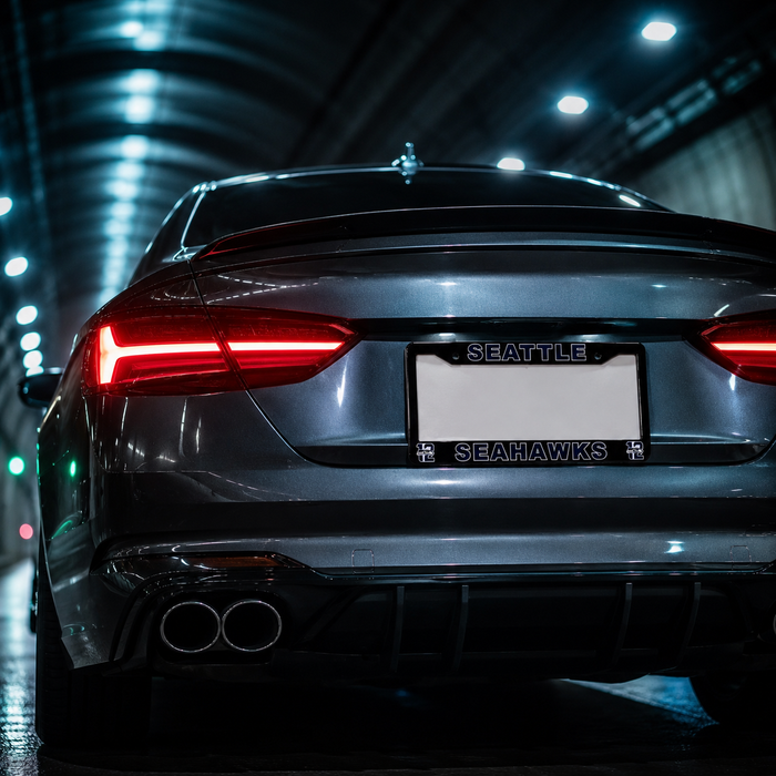 Rear view of a dark gray Seattle Seahawks sports car in a tunnel, featuring custom license plate frames with the team name and logo, illuminated red taillights, and dual exhaust pipes.