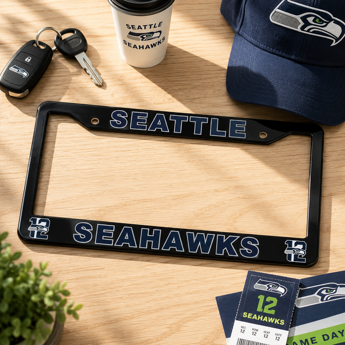 A black Seattle Seahawks license plate frame with navy and white team branding, displayed on a light wood tabletop alongside a Seattle Seahawks coffee cup, keys, a baseball cap, and a ticket stub.