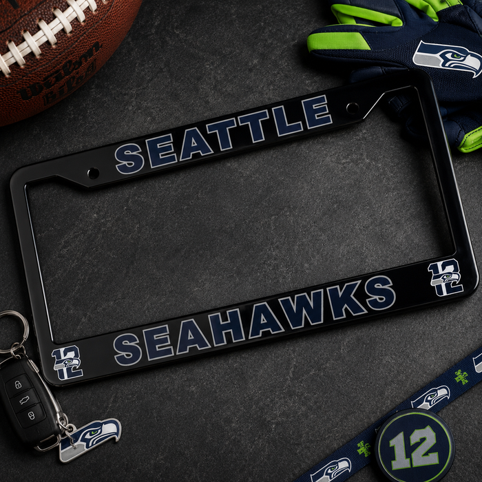 A black Seattle Seahawks license plate frame with white and blue team lettering, displayed on a dark textured surface with Seahawks-themed props including a football, gloves, keychain, and a number 12 patch.