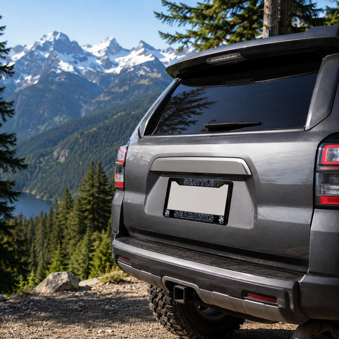 Rear three-quarter view of a gray SUV parked outdoors on a gravel path with a mountain and lake landscape in the background. The license plate frame reads 'SEATTLE' and 'SEAHAWKS'.