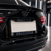 A close-up rear view of a black car showing a custom license plate frame and plate. The frame is black with silver lettering that reads 'SEATTLE' on the top bar and 'SEAHAWKS' on the bottom bar, flanked by team logos.
