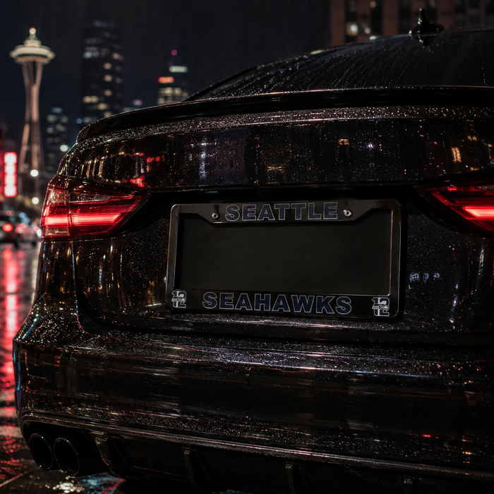 A close-up rear view of a glossy black car on a wet city street at night, featuring a custom license plate frame with 'SEATTLE' at the top and 'SEAHAWKS' at the bottom in blue lettering.
