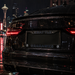 A close-up rear view of a glossy black car on a wet city street at night, featuring a custom license plate frame with 'SEATTLE' at the top and 'SEAHAWKS' at the bottom in blue lettering.