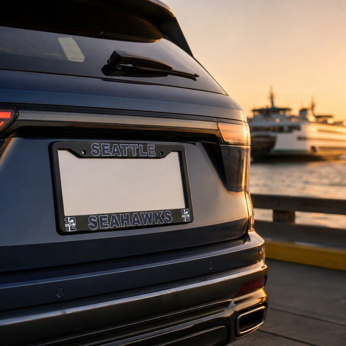 A close-up rear view of a dark SUV parked outdoors near a waterfront at sunset, showing a custom license plate frame with the words "SEATTLE" at the top and "SEAHAWKS" at the bottom, plus Seahawks logos in the corners.