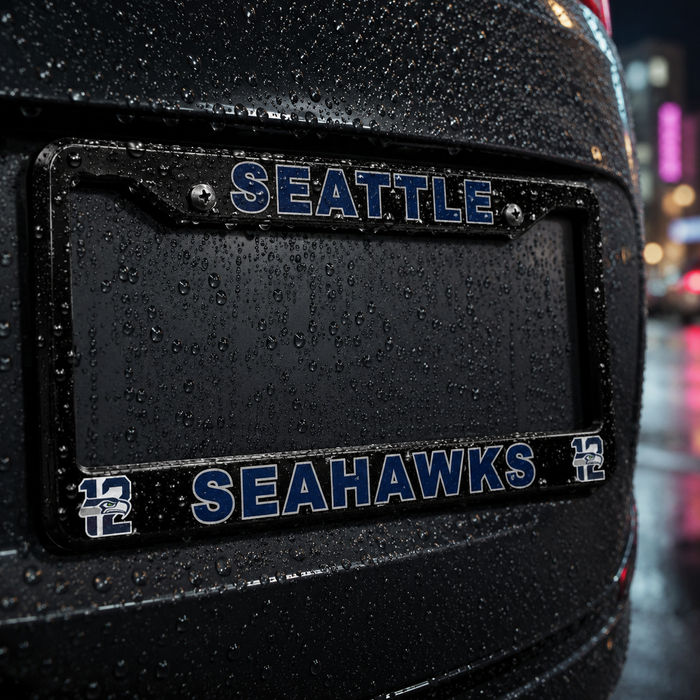 A close-up of a black license plate frame on the rear of a black vehicle, with 'SEATTLE' at the top and 'SEAHAWKS' at the bottom in blue letters outlined in white, shown in rain at night with blurred city lights in the background.
