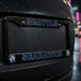 A close-up of a black license plate frame on the rear of a black vehicle, with 'SEATTLE' at the top and 'SEAHAWKS' at the bottom in blue letters outlined in white, shown in rain at night with blurred city lights in the background.