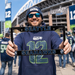A man wearing a Seattle Seahawks beanie and navy hoodie holds up a black license plate frame featuring the team name 'SEATTLE' on top and 'SEAHAWKS' on the bottom, with the Seahawks logo on each corner. The background shows a busy outdoor stadium scene with other fans.