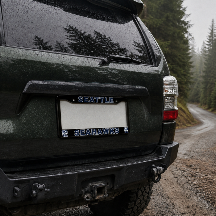 A close-up rear view of a dark green SUV parked on a forest road, showing a custom license plate frame with the word 'SEATTLE' at the top and 'SEAHAWKS' with small hawk logos on the bottom.