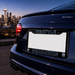 A close-up rear view of a dark blue car with a custom license plate frame featuring 'SEATTLE' at the top and 'SEAHAWKS' at the bottom in Seattle Seahawks team colors, with a blurred city skyline in the background.