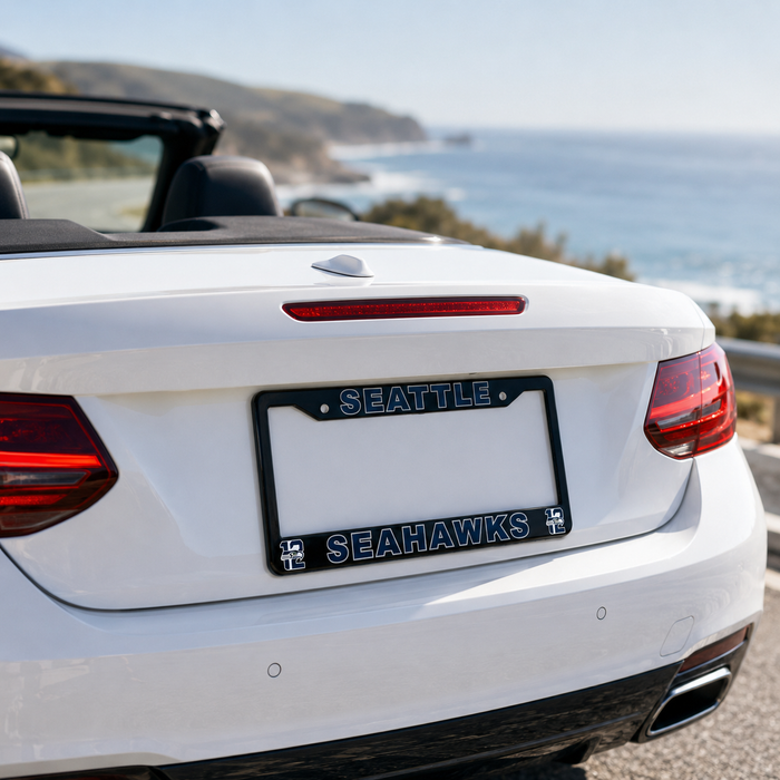 A close-up rear view of a white convertible car on a coastal road, showing a black license plate frame with 'SEATTLE' at the top and 'SEAHAWKS' at the bottom in blue and white lettering.