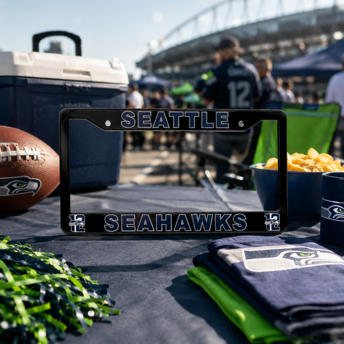 A black Seattle Seahawks license plate frame displayed on a table at a stadium event, surrounded by team merchandise including a football, cooler, chips, and folded towels.