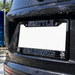 A close-up of a black license plate frame mounted on the rear of a wet black vehicle. The frame has 'SEATTLE' across the top and 'SEAHAWKS' across the bottom, with team logos in the corners and a blank white plate insert.