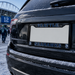 A black license plate frame with 'SEATTLE' on the top bar and 'SEAHAWKS' on the bottom bar, mounted on the rear of a black SUV in the snow near a stadium.