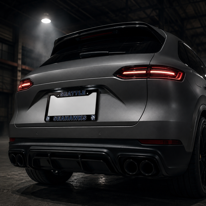 Rear view of a dark gray Porsche SUV in a dim industrial garage setting, featuring a custom license plate frame with 'SEATTLE SEAHAWKS' text and logo.